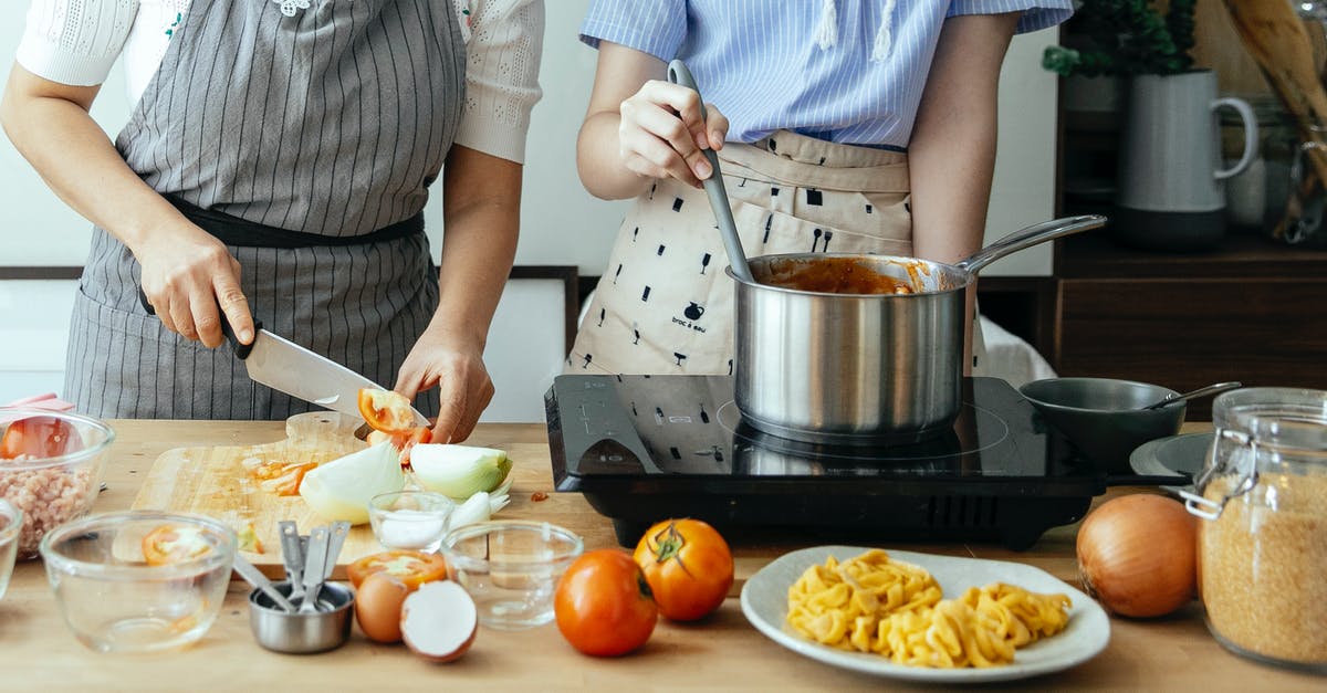 'Caramelization' of tomato sauce in slow cooker - Unrecognizable woman cutting fresh tomato on cutting board while standing at table with ingredients and stove near female cook stirring sauce in pan