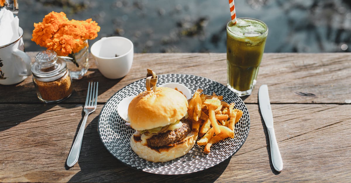 A burger made from pastrami - Plate with appetizing hamburger and french fries placed on lumber table near glass of green drink in outdoor cafe