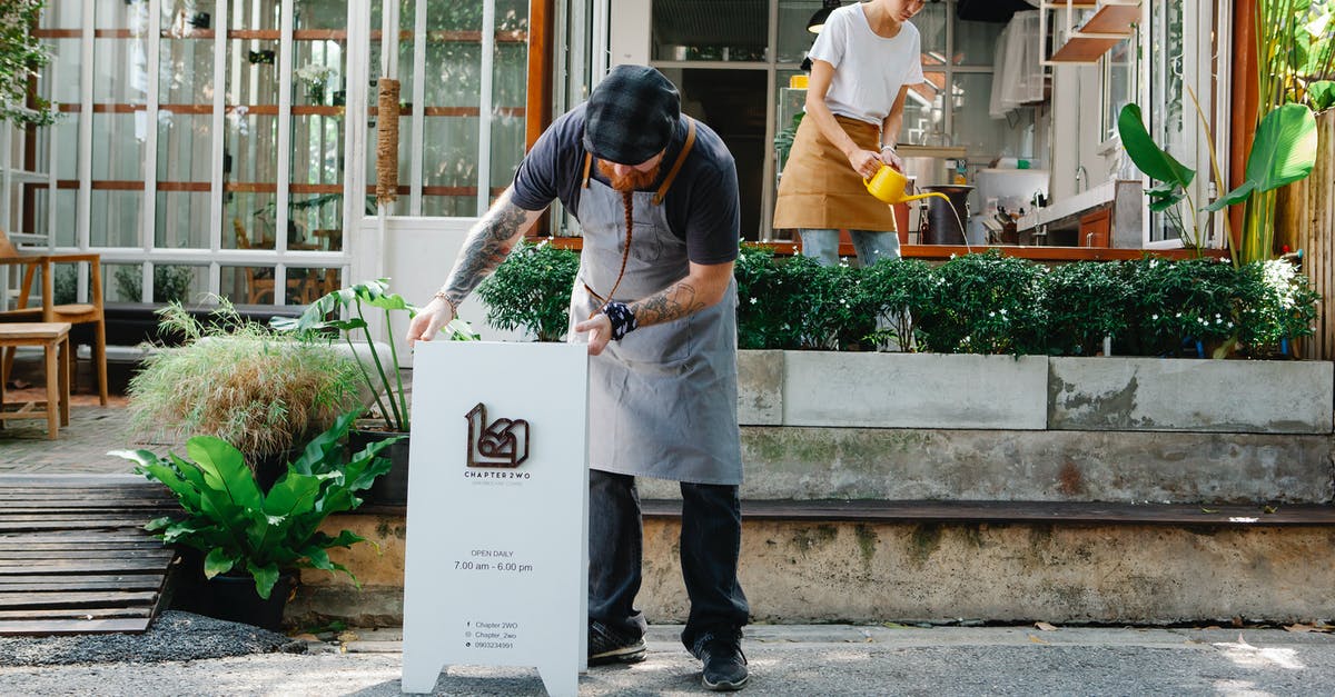 A salad chopper I can put in the dishwasher? - Couple of coworkers in aprons and casual clothes watering plants and putting signboard near cafeteria in daytime