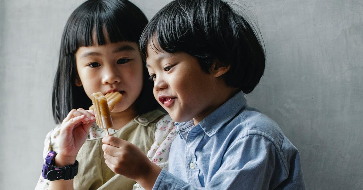 Accidentally beat sugar and butter together - Adorable little Asian siblings with dark hair eating sweet yummy ice pop while standing near gray wall