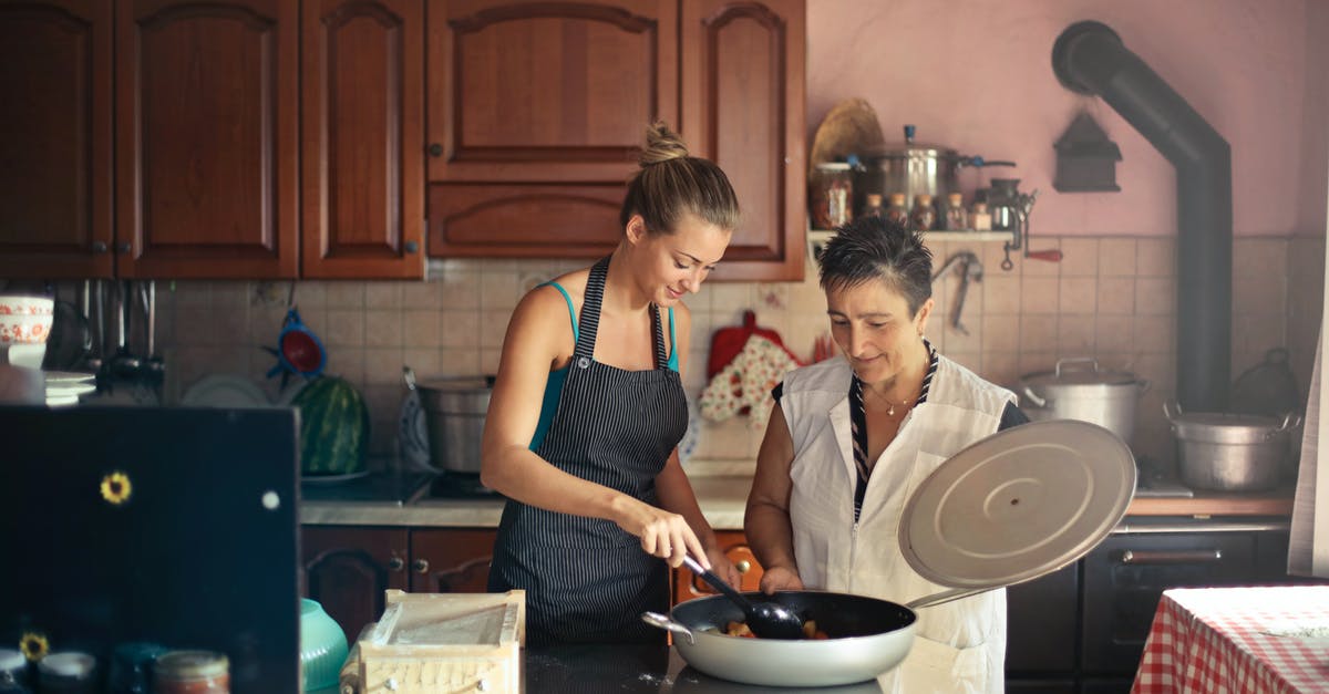 Adding uncooked rice to a slow-cooked stir fry - Daughter and senior mother standing at table in kitchen and stirring dish in frying pan while preparing food for dinner