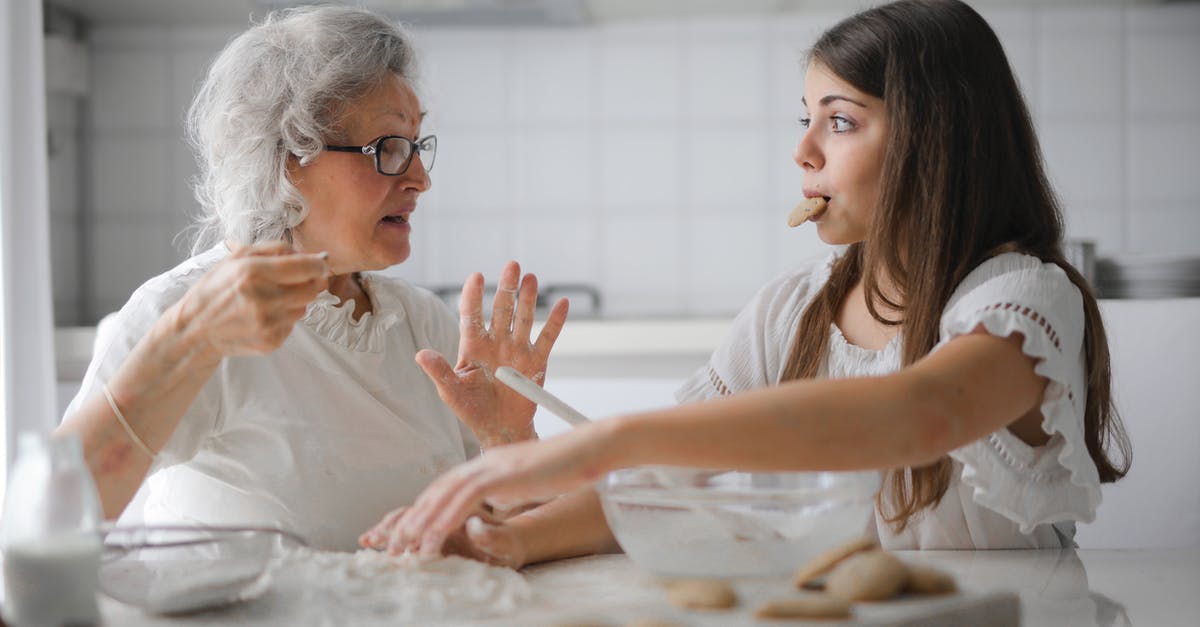 Advice for low temperature cooking - Calm senior woman and teenage girl in casual clothes looking at each other and talking while eating cookies and cooking pastry in contemporary kitchen at home