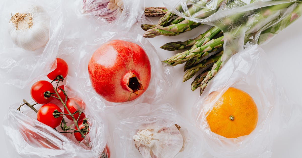 After few day of plastick bag packing of Tomato sauce increase the size of it and produce gas in plastic bag - Top view of pomegranate in center surrounded by bundle of raw asparagus with orange and bunch of tomatoes put near heads of garlic in plastic bags on white surface