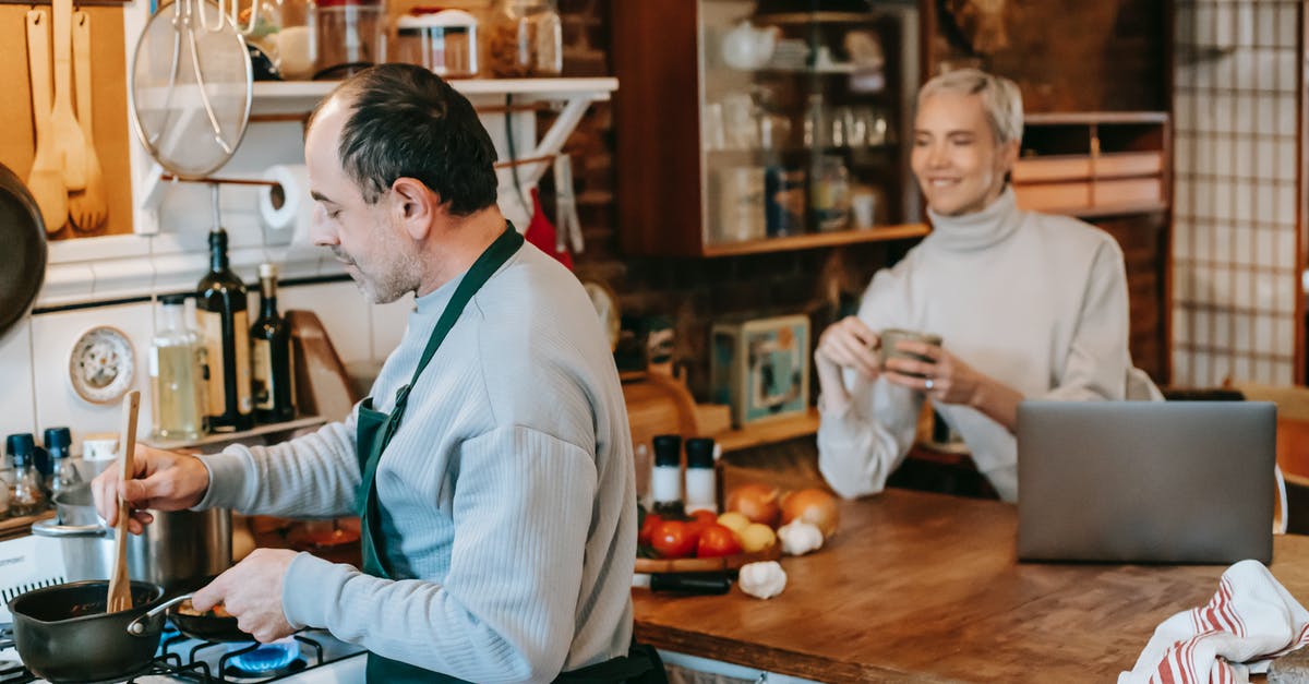 Avoiding over-mixing when using a stand mixer - Focused male preparing dish on gas stove in kitchen with smiling wife having drink and using laptop