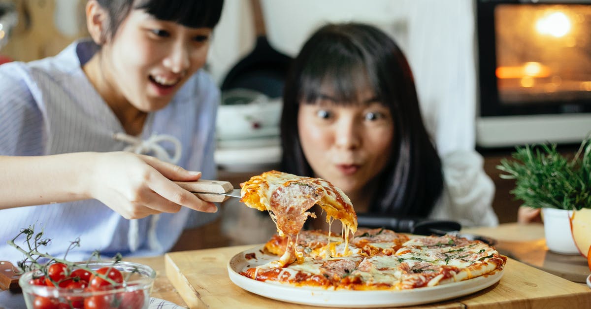 Baking pizza without an oven - Crop delightful Asian ladies smiling while cutting piece of delicious homemade pizza with stretched cheese on cutting board in kitchen