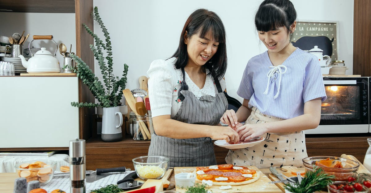 Baking pizza without an oven - Happy smiling Asian mother and daughter in aprons putting ingredients on dough while cooking pizza together in kitchen