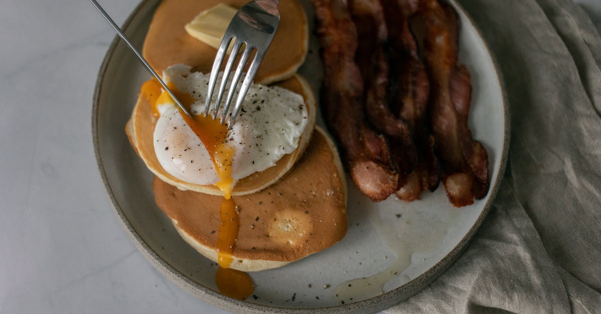 Beaten and poached eggs - From above of unrecognizable person cutting poached egg served on pancakes with roasted bacon during breakfast in kitchen