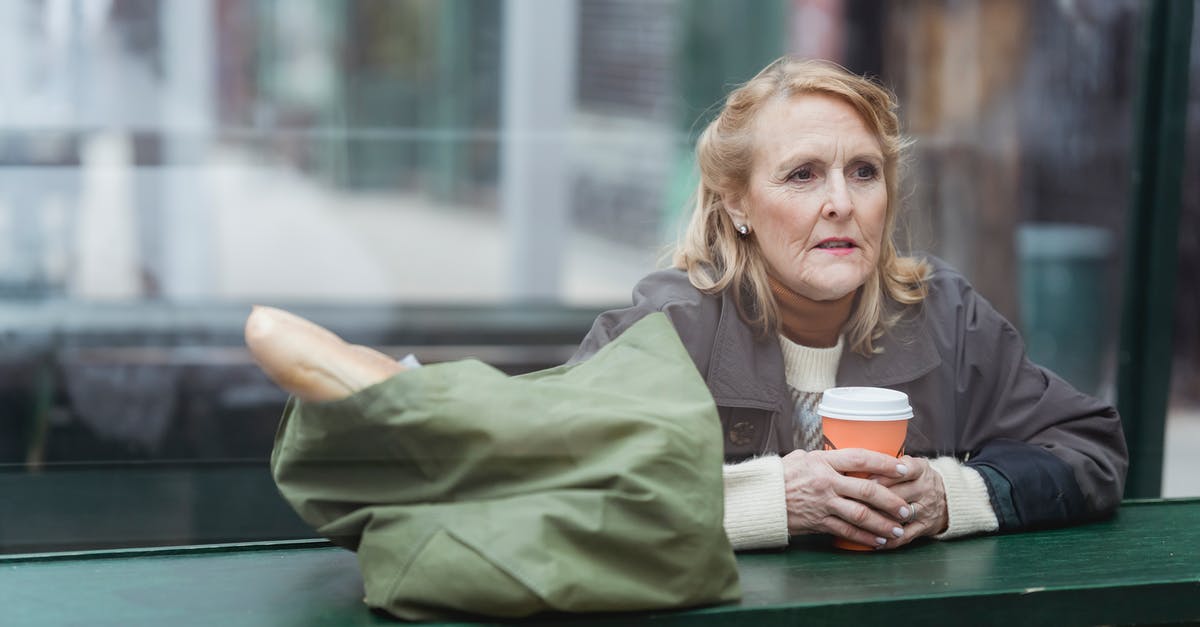 Bread going stale in enamel breadbasket - Contemplative senior female with disposable cup of hot drink and baguette in bag looking away at cafe counter