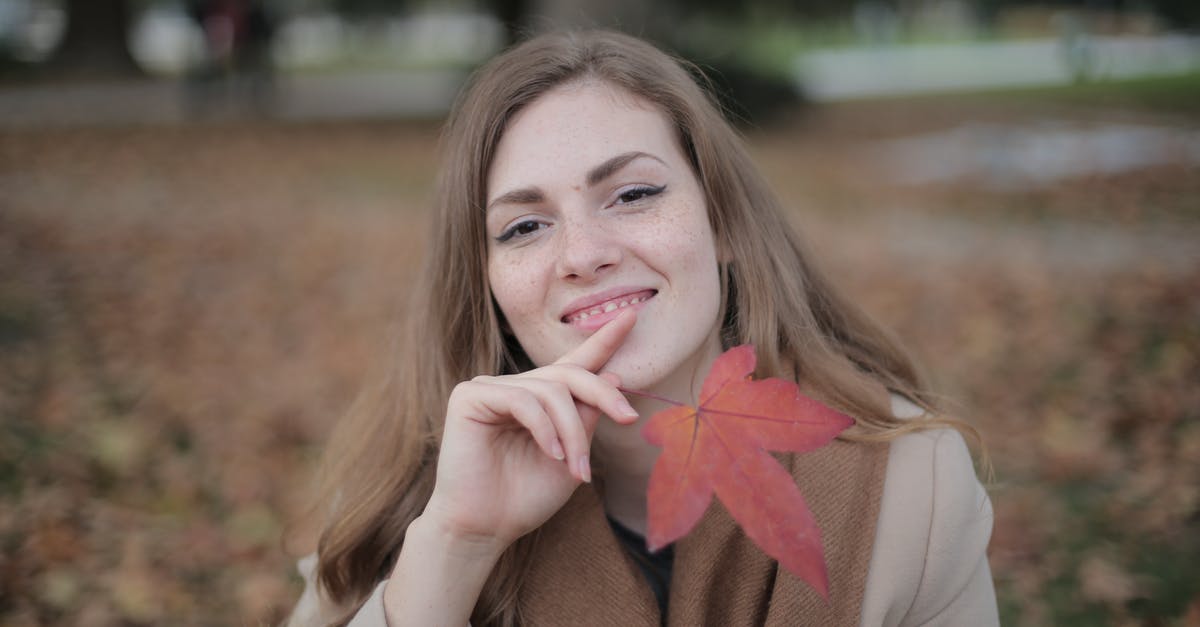 Brown Blob in natural maple syrup? - Attractive young lady in warm clothes and scarf with red maple leaf touching chin and smiling at camera against blurred environment in autumn park