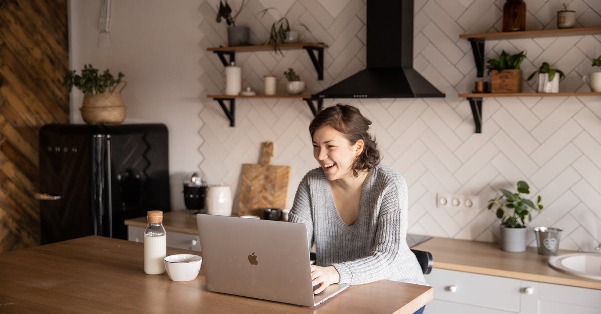 Burnt milk uses - Young female in gray sweater laughing while sitting at wooden table with laptop and milk bottle and having rest watching video during free time