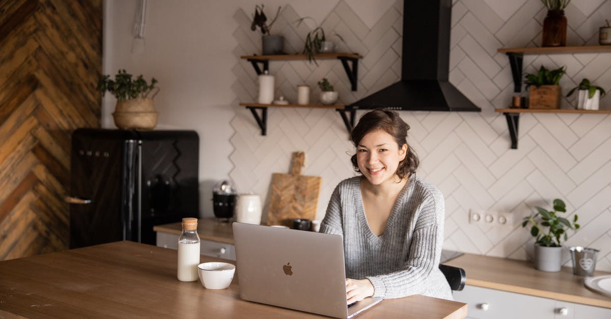 Burnt milk uses - Cheerful female in casual clothes sitting at table with laptop and bottle of milk while browsing internet on laptop during free time at home and smiling looking at camera