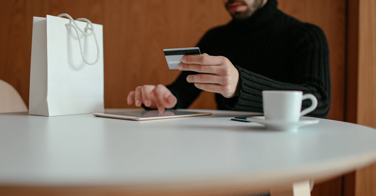 Buying chocolate transfer sheet - Crop focused bearded young man using tablet while doing online purchase with credit card sitting at table in modern cafe with coffee cup and shopping bag