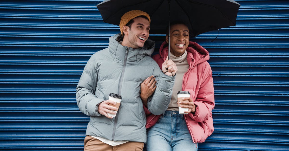 Caffeine content of cold-brewed coffee: higher or lower than hot-brewed? - Happy multiracial couple with umbrella in warm outerwear laughing while standing on street with takeaway beverages in cold rainy weather