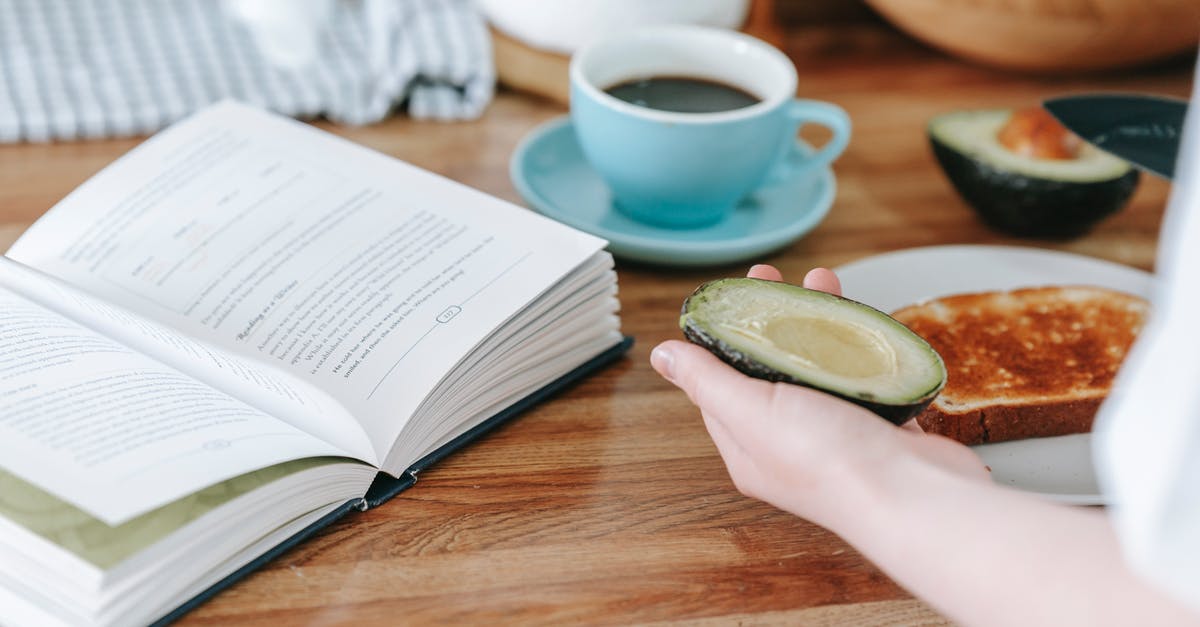 Calculating Nutrition Information for Commercial Food Labeling - Crop anonymous person sitting at table and reading book while having coffee and making toast with avocado for breakfast