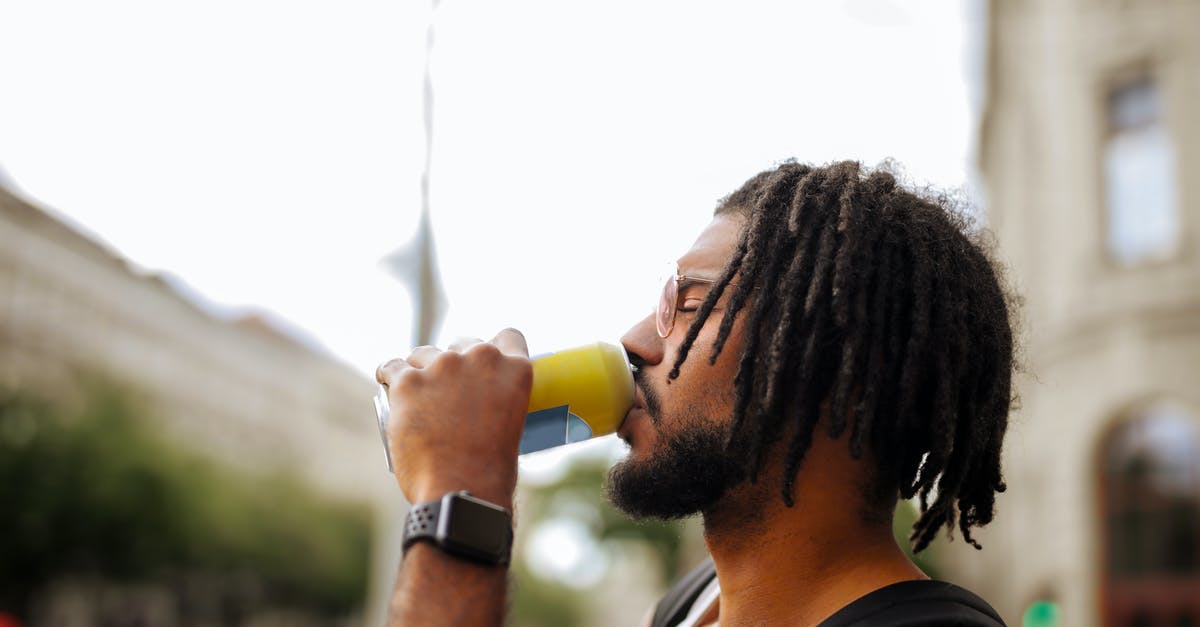 Can I cool my toffee in the fridge? - Side view of adult Hispanic guy with dreadlocks in sunglasses and casual clothes with backpack and smart watch drinking yummy beverage from vivid yellow can while standing with eyes closed on street in downtown