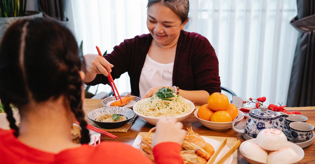 Chinese rice vermicelli tastes of chemicals - Cheerful ethnic grandma with cooked prawn against anonymous female teen at table with assorted tasty dishes at home