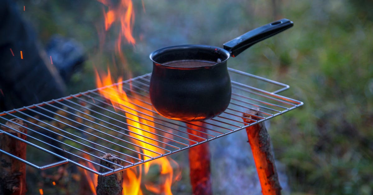 choosing a saucepan material for creams and caramels - Metal saucepan with beverage on metal rack above blazing bonfire in woods in daytime