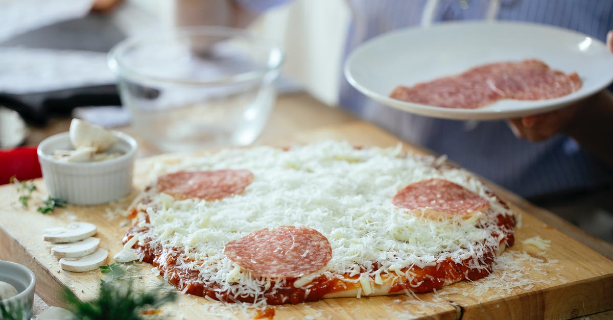 Coating meat in flour before browning, bad idea? - Crop anonymous person standing near table with uncooked homemade pizza and adding slices of salami while preparing in kitchen at daytime