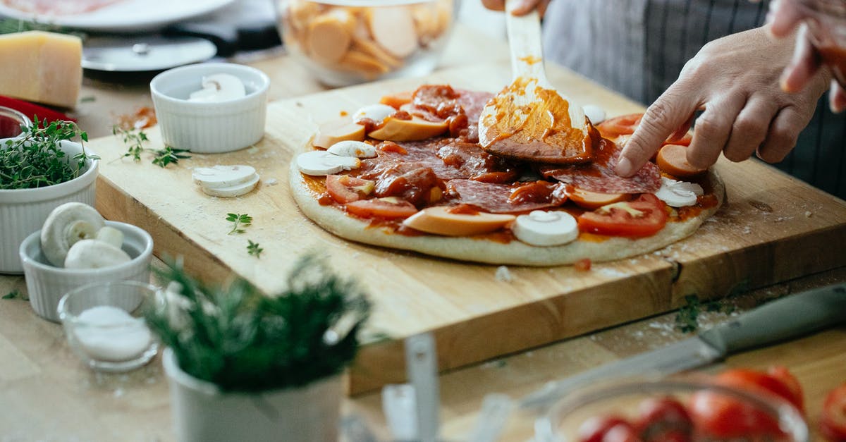 Cooked or raw mushrooms on pizza? - Unrecognizable chef spreading sauce on pizza with tomatoes mushrooms and salami placed on wooden cutting board on table with ingredients