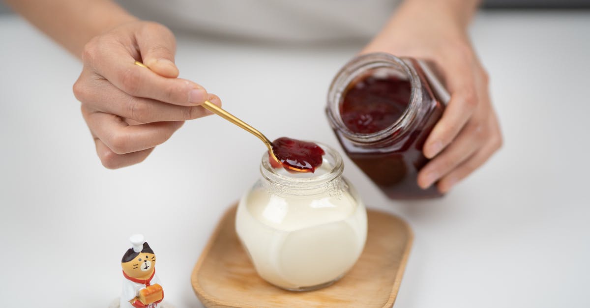 Cooking protein marinated in yogurt - From above crop faceless person adding berry jam to plain organic yogurt in glass jar placed on wooden saucer near tiny cat statuette