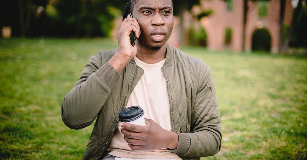 Could it be bad black olive that I've eaten? - Young African American male in casual clothes with cup of coffee to go in hand talking on mobile with expression of displeasure and misunderstanding on face while sitting in park