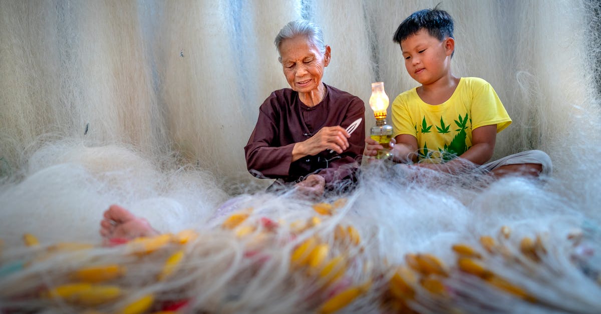 Couldn't make pesto in a blender - Focused elderly barefoot ethnic grandma with needle mending fishing net against boy with kerosene lamp