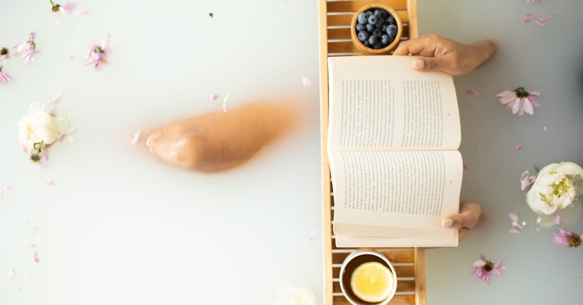 Creating an eggwash with water vs. milk - Top view of crop unrecognizable lady in white water in bathtub with fresh colorful flower petals with wooden tray with cup of tea with lemon and blueberries while reading book