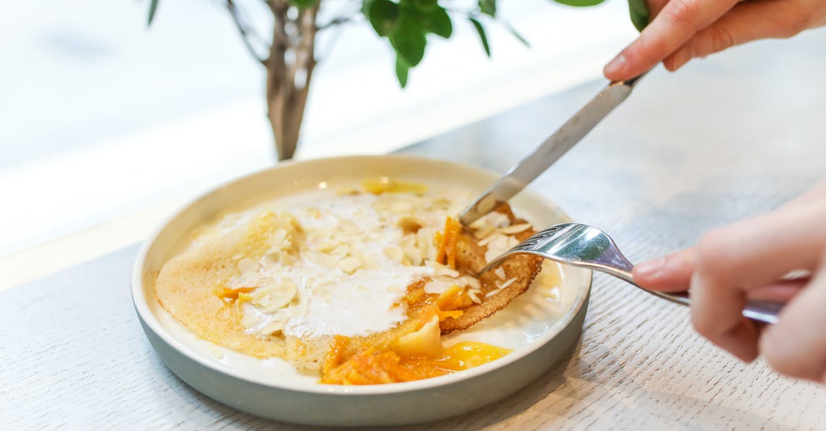 Crepe vs Pancake - Unrecognizable crop person eating sweet tasty pancakes served on white plate with fork and knife while sitting at wooden table in light room