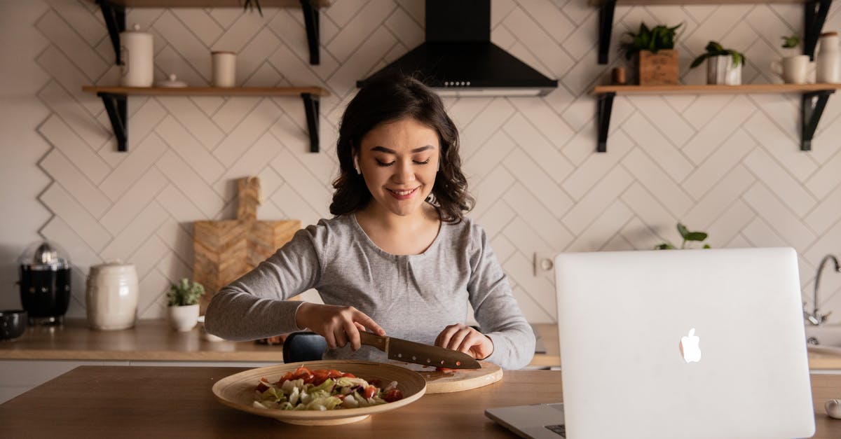 Culinary uses for honeycomb? - Cheerful ethnic female cutting fresh vegetables on cutting board while sitting at wooden table in kitchen with open portable computer in apartment