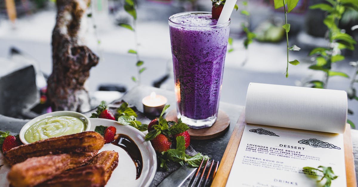 Culinary uses for lavender - From above of glass of lavender creme brulee garnished with strawberry and plate of churro with avocado sauce placed on table with knife and fork and menu and candle against window decorated with plants