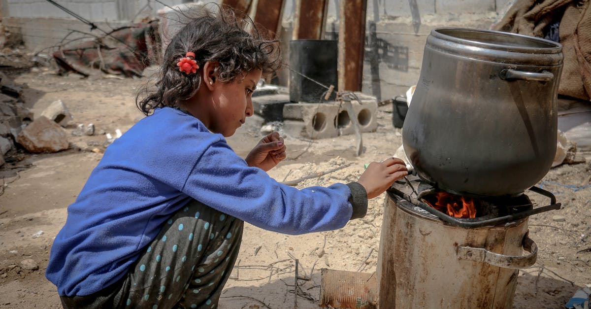 Damage to pan bottom - Side view of ethnic kid sitting near furnace with metal pan placed on fire in daylight in poor destroyed district