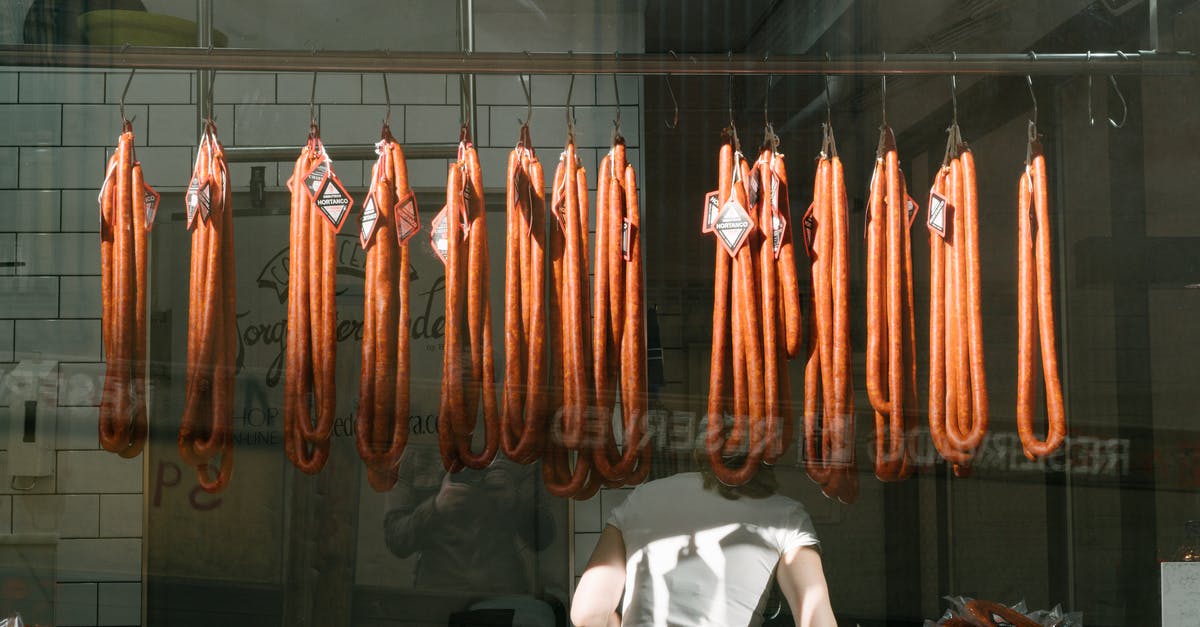 Dealing with a hank of natural sausage casings - Through glass back view of unrecognizable worker in white T shirt at meat preserving factory with long sausages hanging from metal rail in tiled room