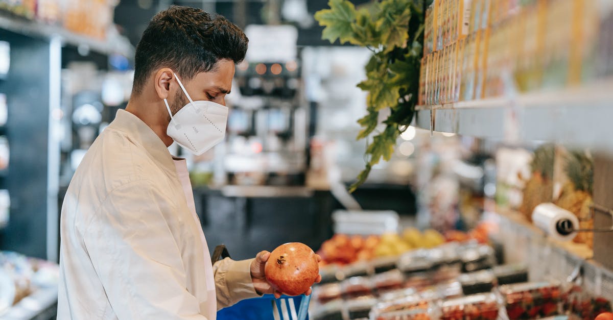 Dehydrating fruit/veg, temperature, and safety - Side view of young Hispanic man in protective mask choosing fresh pomegranate from stall in supermarket during coronavirus pandemic