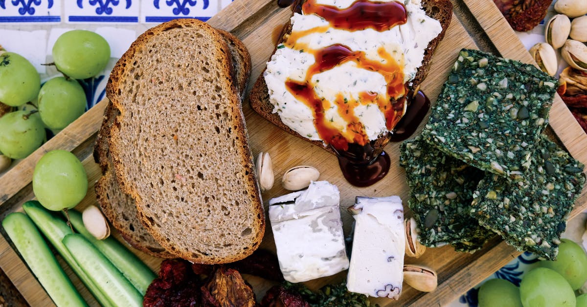 Difference between French bread and Japanese bread - Top view of wooden cutting board with fresh cucumber slices and black bread loafs with soft tofu cheese near dry seaweed and crunchy pistachios near pineapple guava