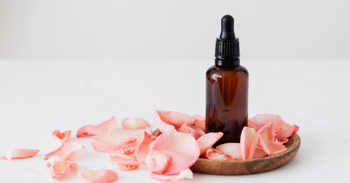 Distributing small amounts of oil around a non-stick pan - Gentle pale pink wavy rose petals placed on small round wooden plate and table near dark glass essence flask on white background