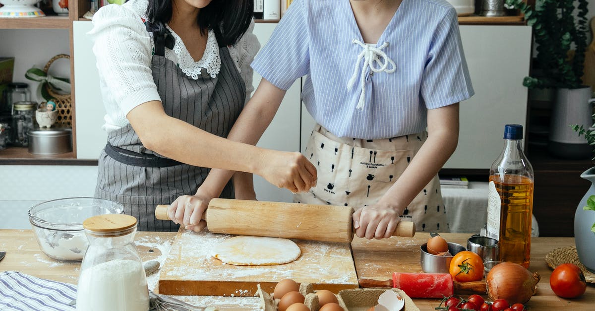 Do onions help to sweeten tomato sauce? - Crop anonymous mother standing near table with daughter while cooking in aprons with flour and eggs while rolling pastry at home