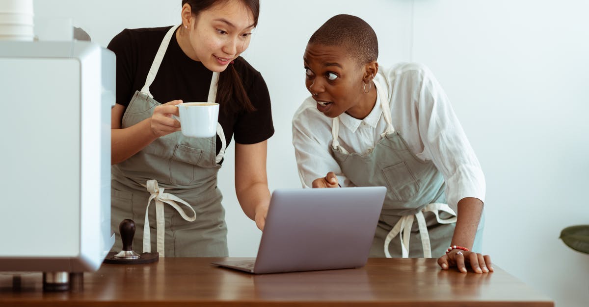 Do professional cooks use measuring cups and spoons? - Cheerful surprised diverse colleagues in aprons using modern netbook and sharing business ideas while gathering at table with cup of drink in light cozy kitchen