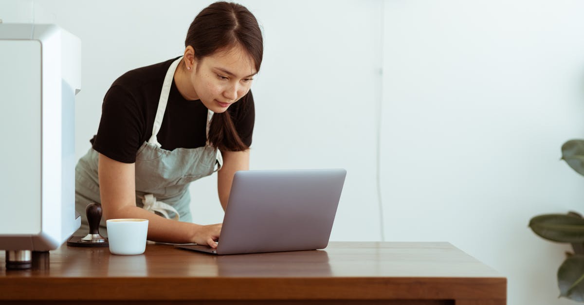 Do professional cooks use measuring cups and spoons? - Concentrated young Asian lady in casual black shirt and apron with cup of hot drink watching cooking tutorials via modern netbook on wooden table against white wall