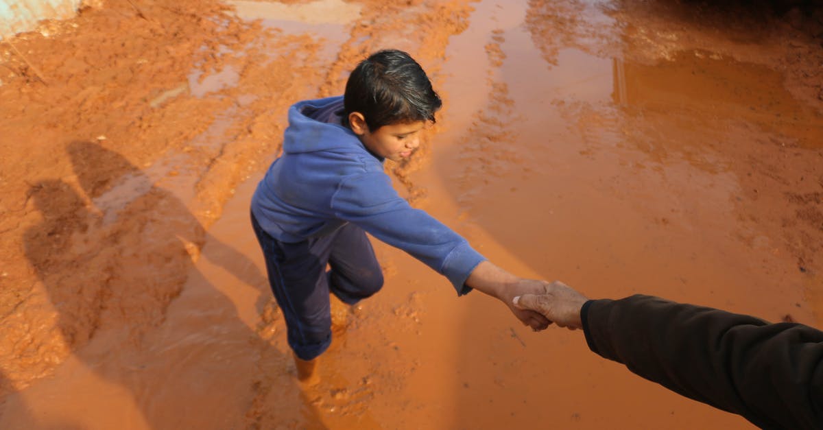 Do sprats need to be gutted? - High angle of crop person holding hands with ethnic boy stuck in dirty puddle in poor village