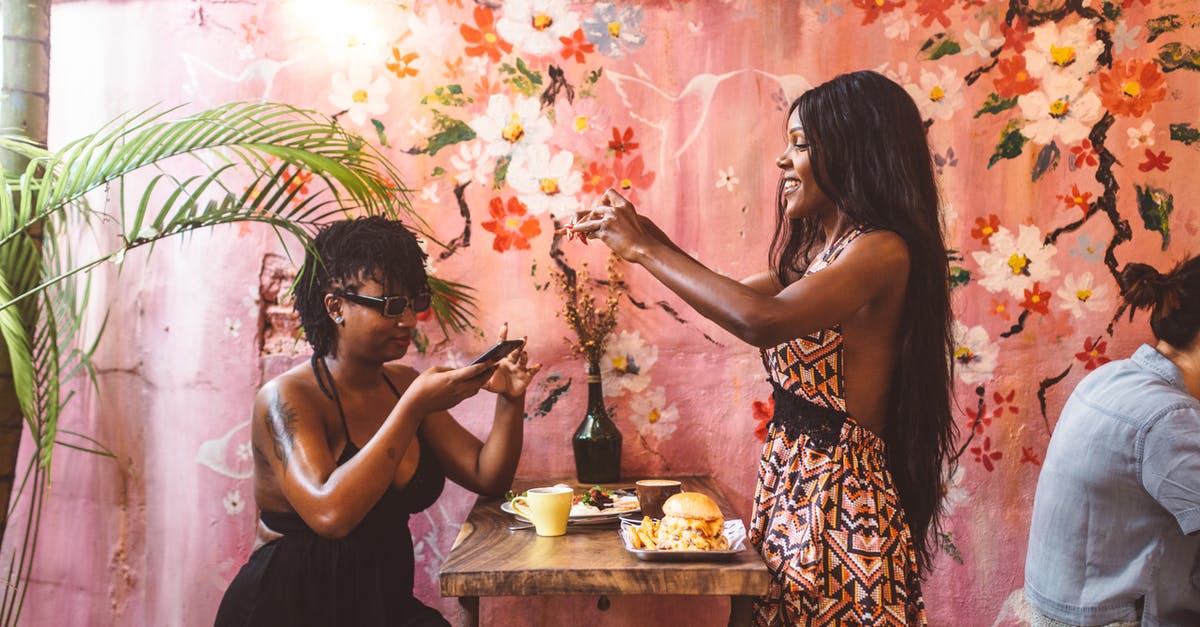 Does "American" Food exist around the world? - Side view happy African American females in stylish dresses taking photos on smartphones of freshly cooked food while having lunch in cozy cafe against painted wall