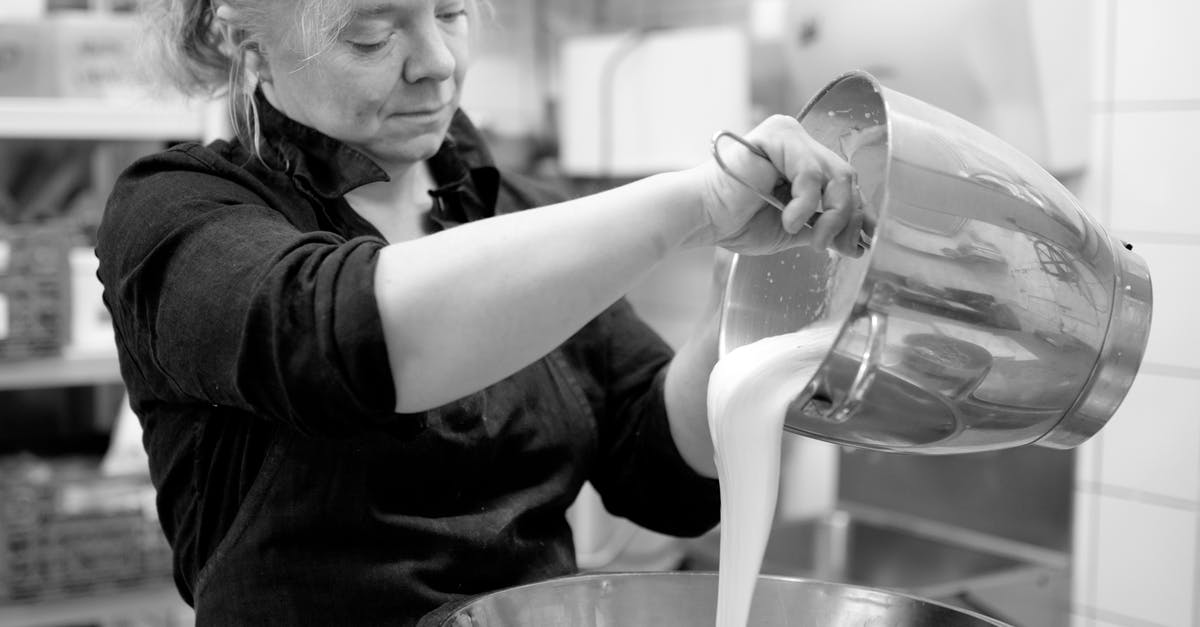 Fatty liquid vs non fatty liquid in dough - Black and white of focused adult female chef in black wear with hair tied up pouring white liquid eatable mass in big glistening metallic dish