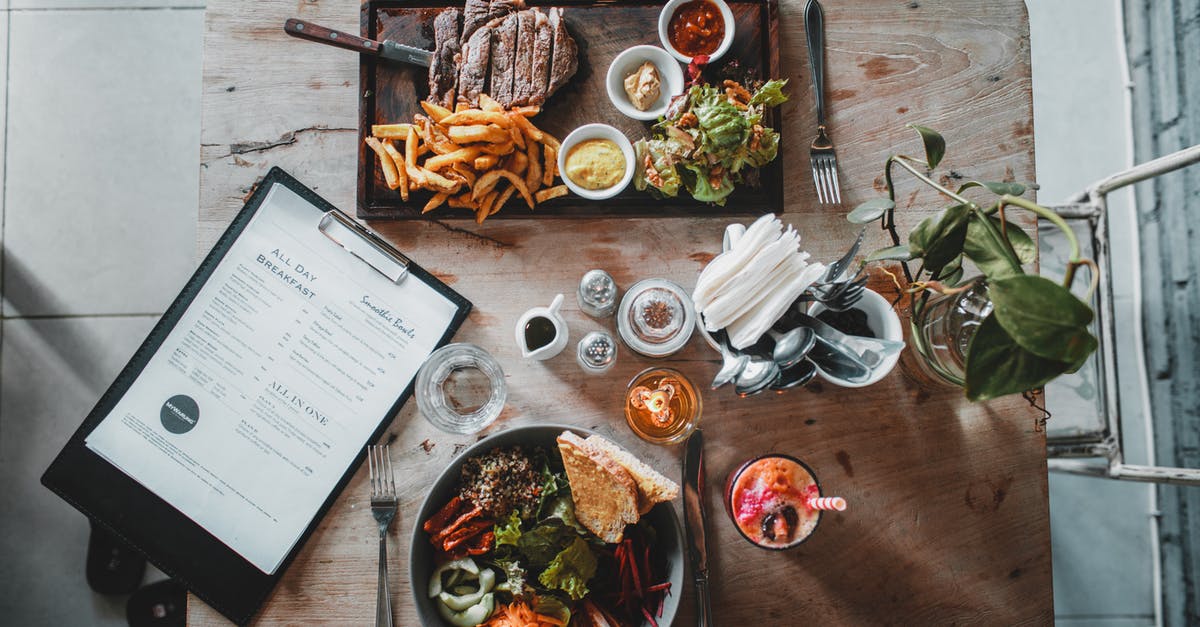 Faulty recipes: omitted ingredient or unused ingredient - Top view of wooden table with salad bowl and fresh drink arranged with tray of appetizing steak and french fries near menu in cozy cafe