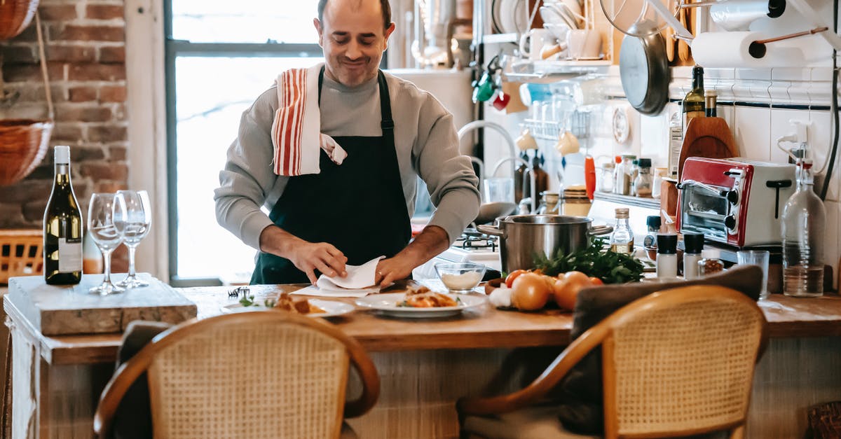 Flambéing wine to cook off alcohol - Positive middle aged male cooking food in bright room near counter with plates with pasta near onion and herbs near kitchenware and wine bottle near glasses and chairs near window