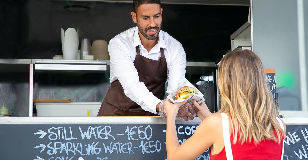 Food industry: where does buttermilk go? - Positive ethnic cook in apron standing at counter in food truck and giving delicious hamburger to anonymous woman customer in daytime