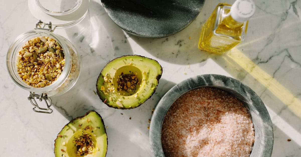 Fruit Oil Extraction - Top view of tasty halved avocado sprinkled with various seeds placed near pink salt with seed mixture in glass pot and other dressing ingredients arranged on white marble table