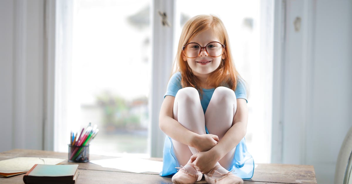 Ginger precipitation - Joyful red haired schoolgirl in blue dress and ballet shoes smiling at camera while sitting on rustic wooden table hugging knees beside school supplies against big window at home