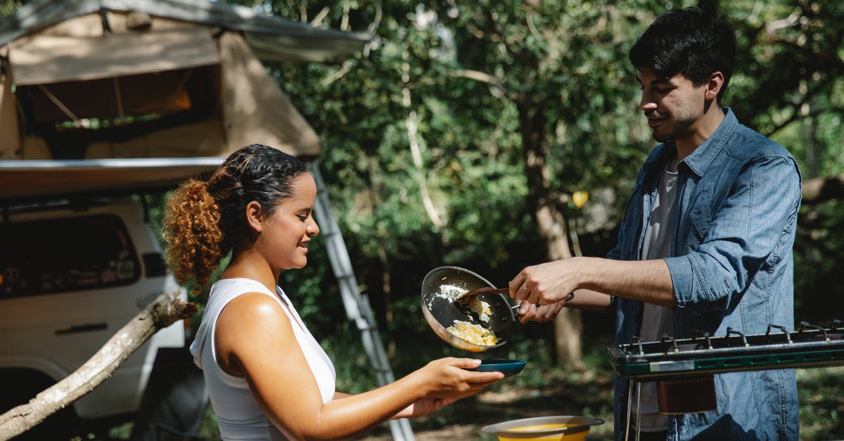 Gravy with Scrambled Eggs :: What went wrong? - Side view of cheerful young multiracial couple in casual clothes putting scrambled eggs from skillet on plate standing near metal stove in camp