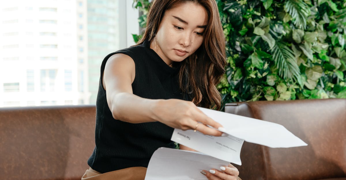 Grilling burgers: flip once, or keep flipping? - Focused Asian female turning pages of document while sitting on sofa during paperwork in modern workspace with green deciduous plant