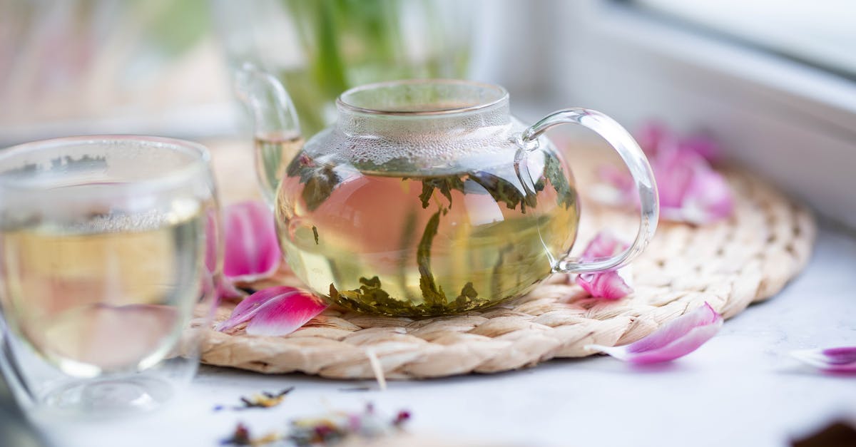 Grinding loose Green Tea for more concentration - Transparent teapot with loose leaf tea placed on wicker mat with petals on white windowsill with glass on blurred background