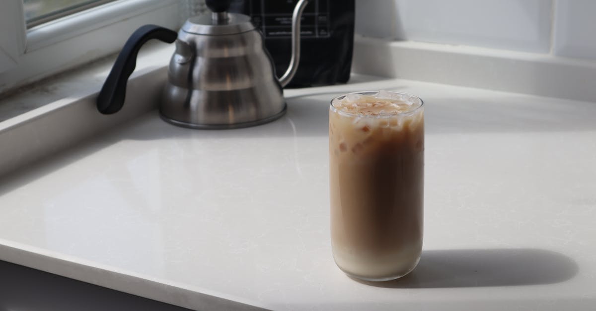 Homemade coconut milk in coffee compared to store bought - High angle glass of delicious iced latte and metal coffee kettle placed on white table in kitchen